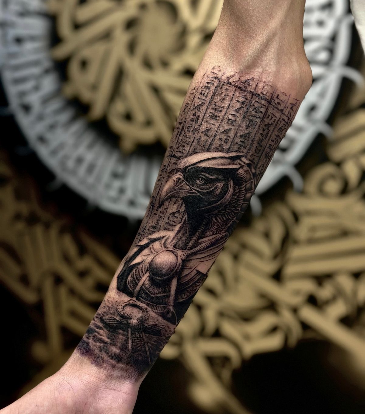 A detailed black and grey forearm tattoo of an Egyptian bird deity and hieroglyphics.