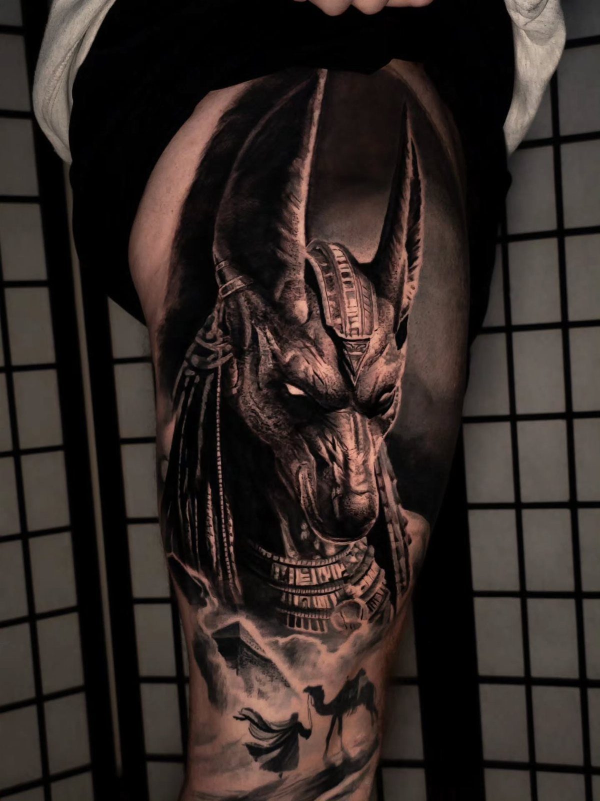 Black and grey tattoo of an Egyptian jackal god with ornate jewelry on a person's thigh.