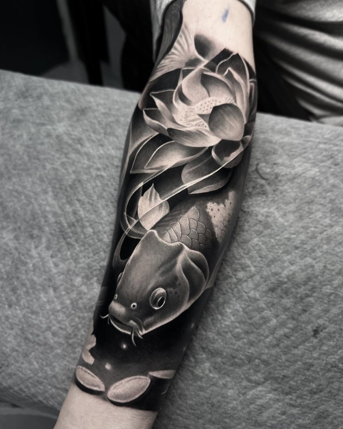 Black and grey tattoo of a koi fish and a lotus flower on a forearm.