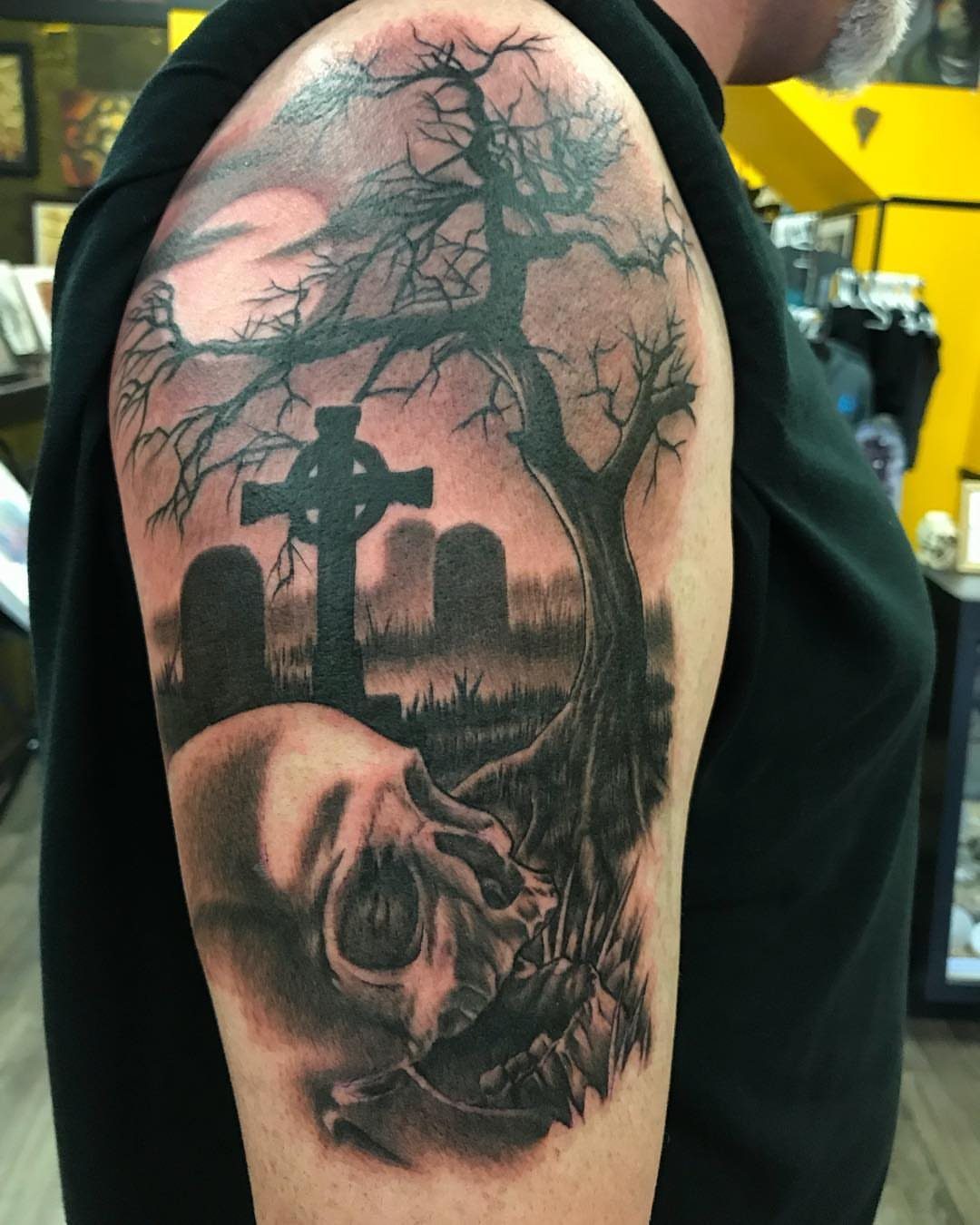 A large black and grey tattoo of a skull and graveyard on a person's shoulder.