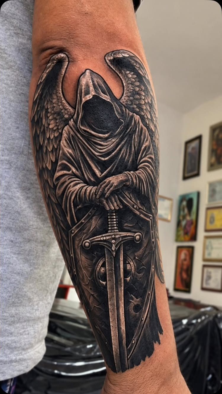 A black and grey realism tattoo of a hooded winged figure holding a sword and shield.