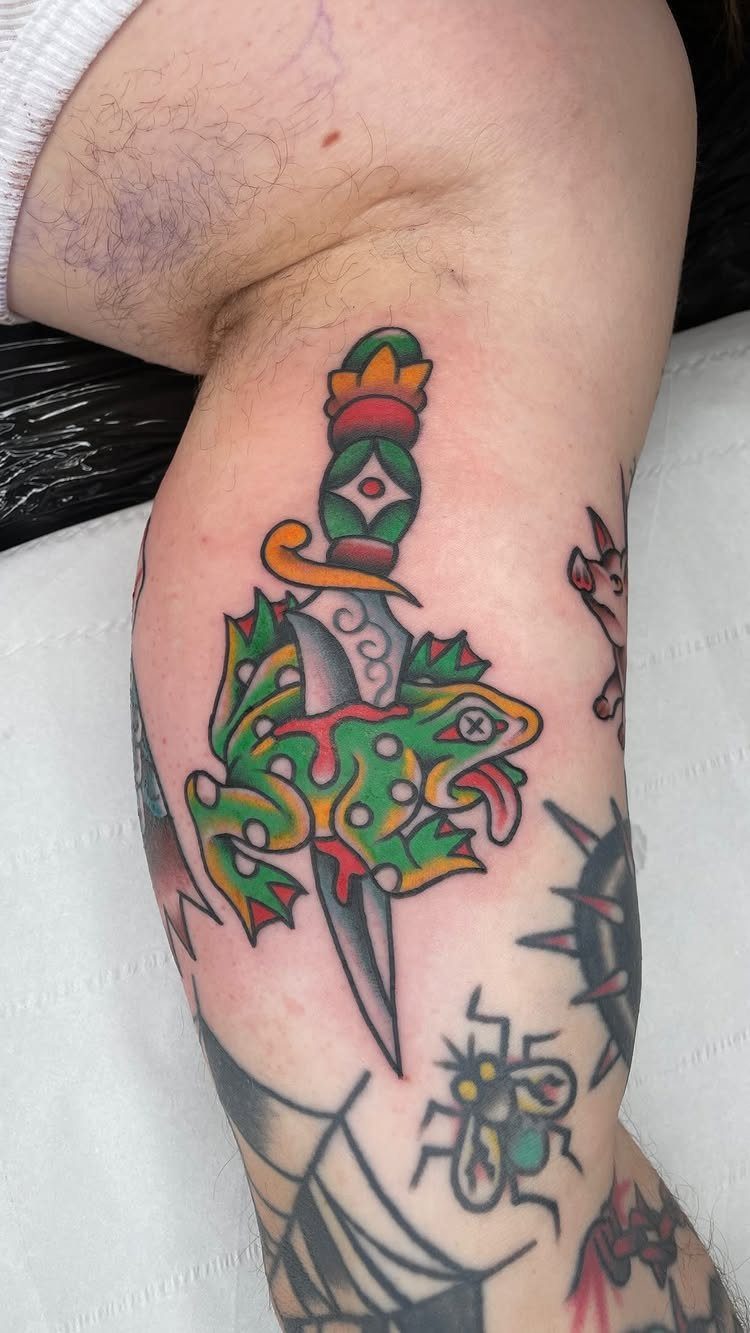 A colorful American traditional tattoo of a dagger piercing a green frog on a person's leg.