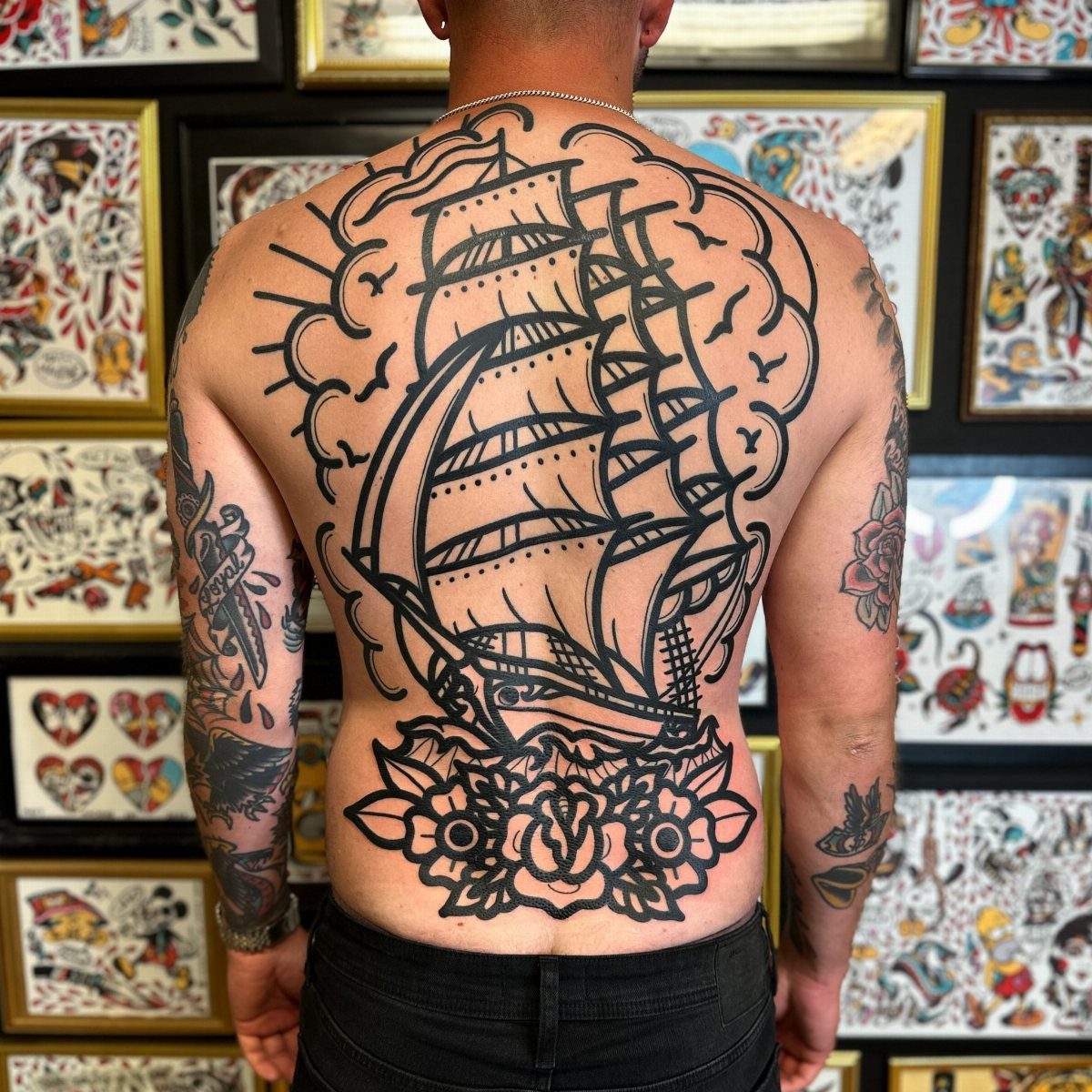 A large black ink sailing ship tattoo covering a person's entire back with floral details.