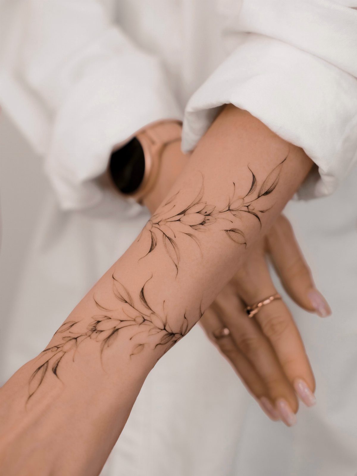 A forearm displaying a delicate black ink tattoo of botanical leaves and floral patterns.