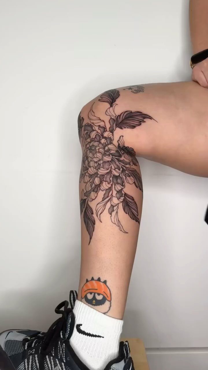 A large blackwork chrysanthemum tattoo with detailed leaves wrapping around a person's leg.