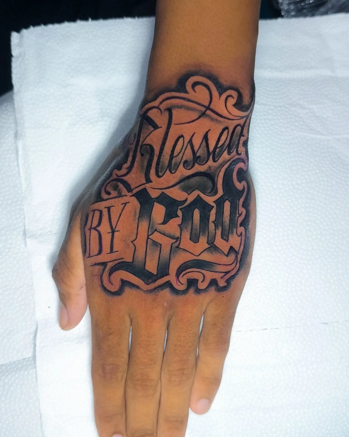 A hand featuring a large black and grey script tattoo that reads Blessed BY GOD.