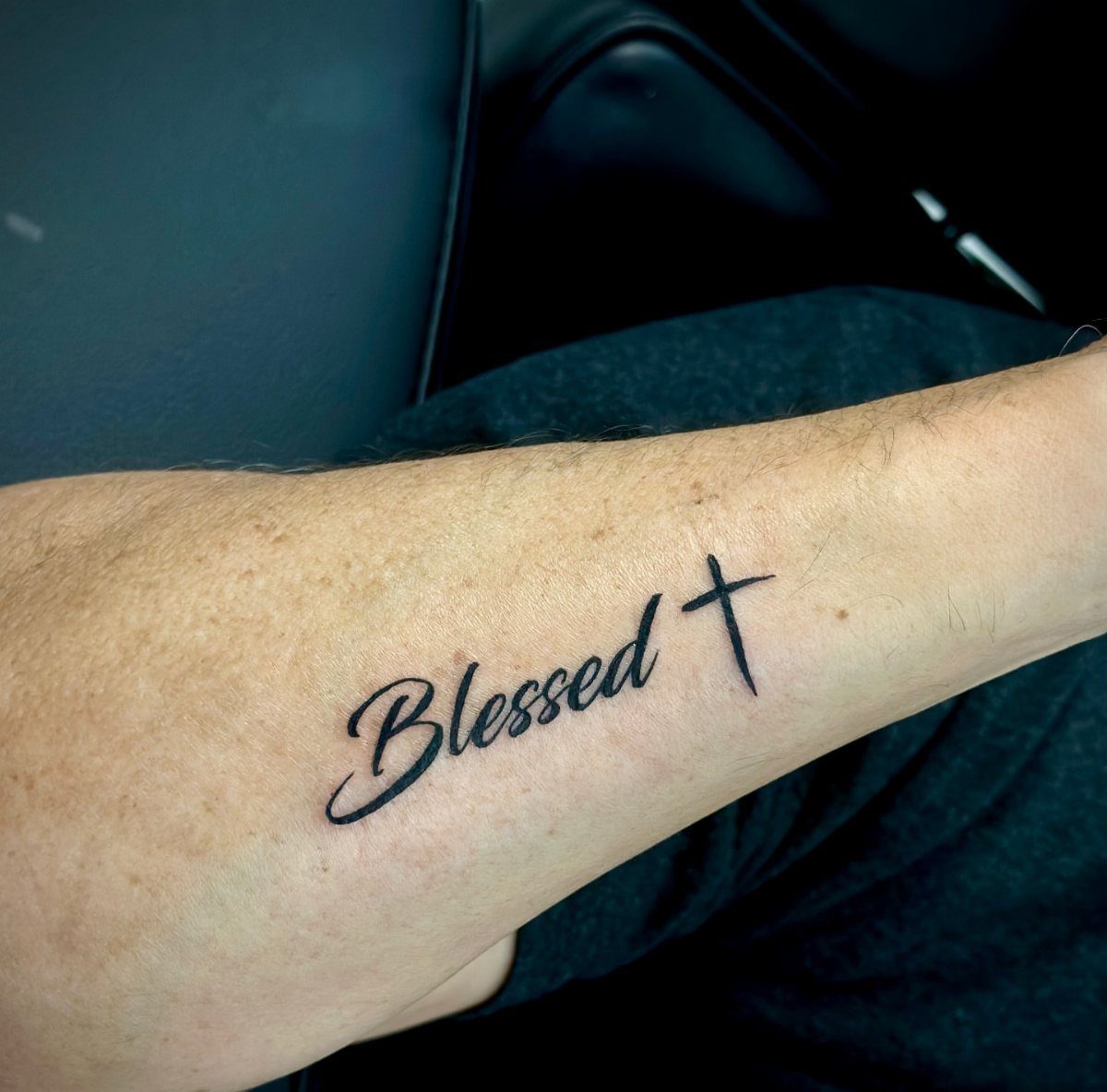 A black ink tattoo on a person's forearm reading Blessed followed by a small cross.