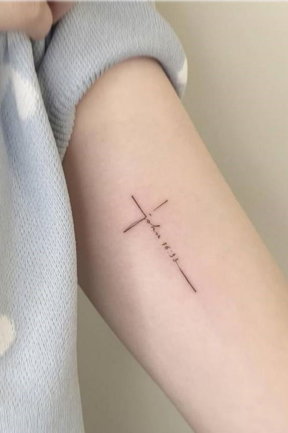 A minimalist fine-line black ink tattoo of a cross with cursive text on an arm.