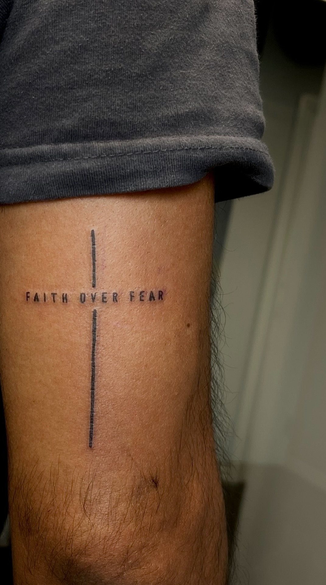 A minimalist black ink tattoo of a cross with the words faith over fear on a leg.