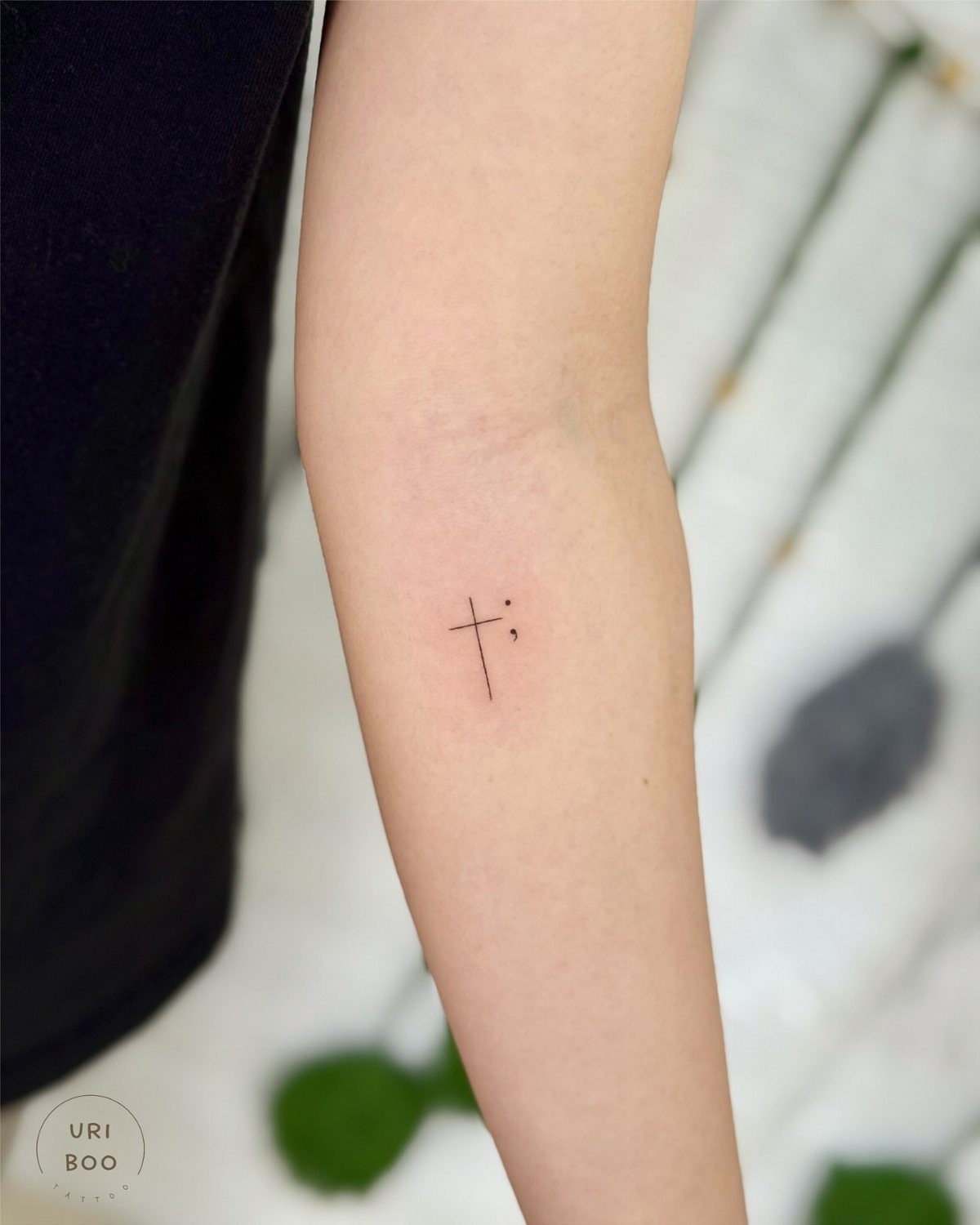 A small fine-line black ink tattoo of a cross and a semicolon on a forearm.