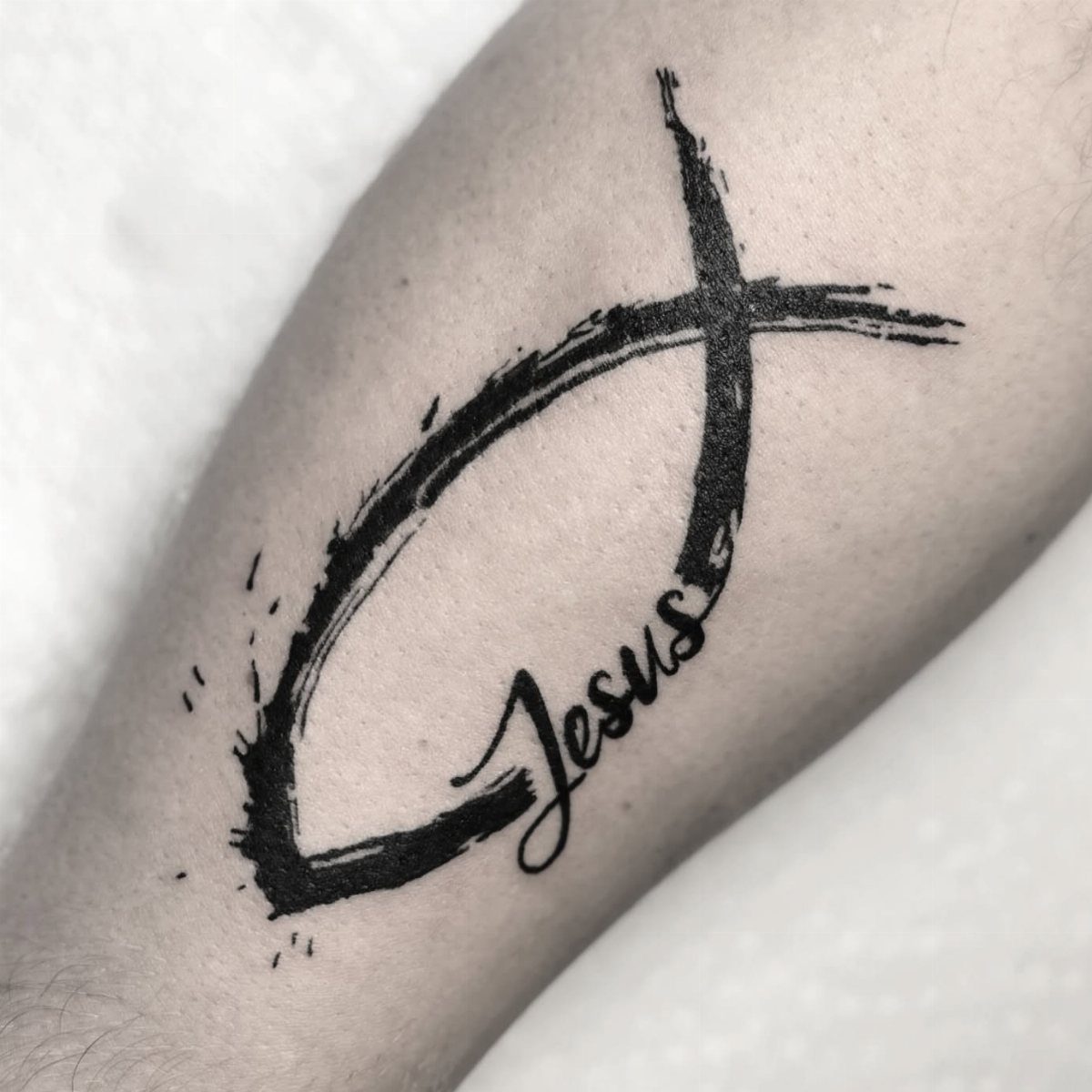 A black ink ichthys fish symbol tattoo with the word Jesus written in cursive script.