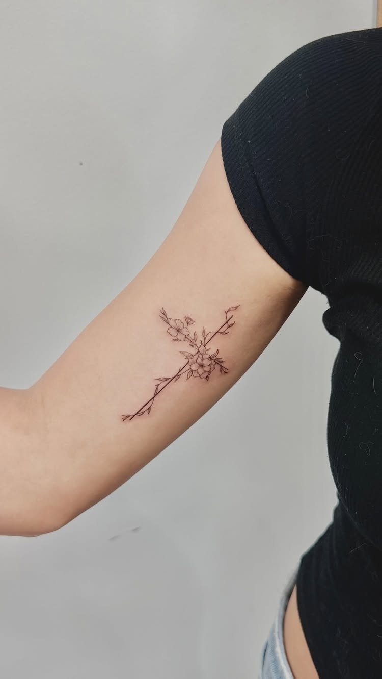 A fine-line black ink tattoo of a floral cross on a person's arm.