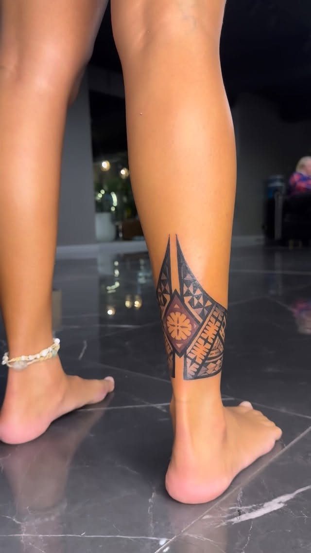 A person's lower legs featuring a large black geometric tribal tattoo wrapped around the ankle.