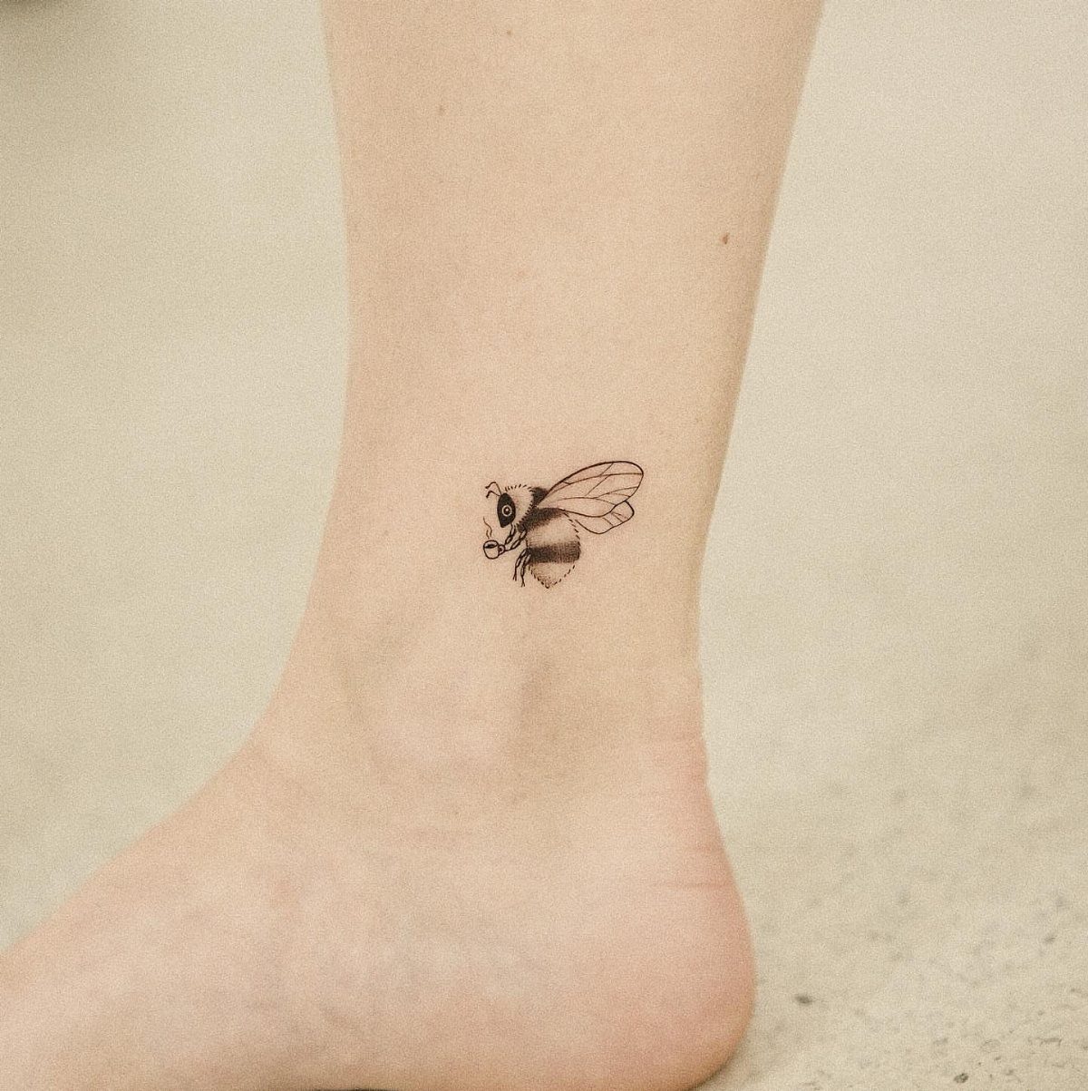 A small black ink bee tattoo on a person's ankle against a neutral background.