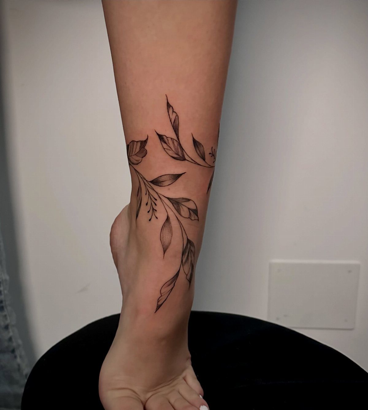 A person's ankle featuring a delicate black ink botanical leaf tattoo wrapping around the skin.