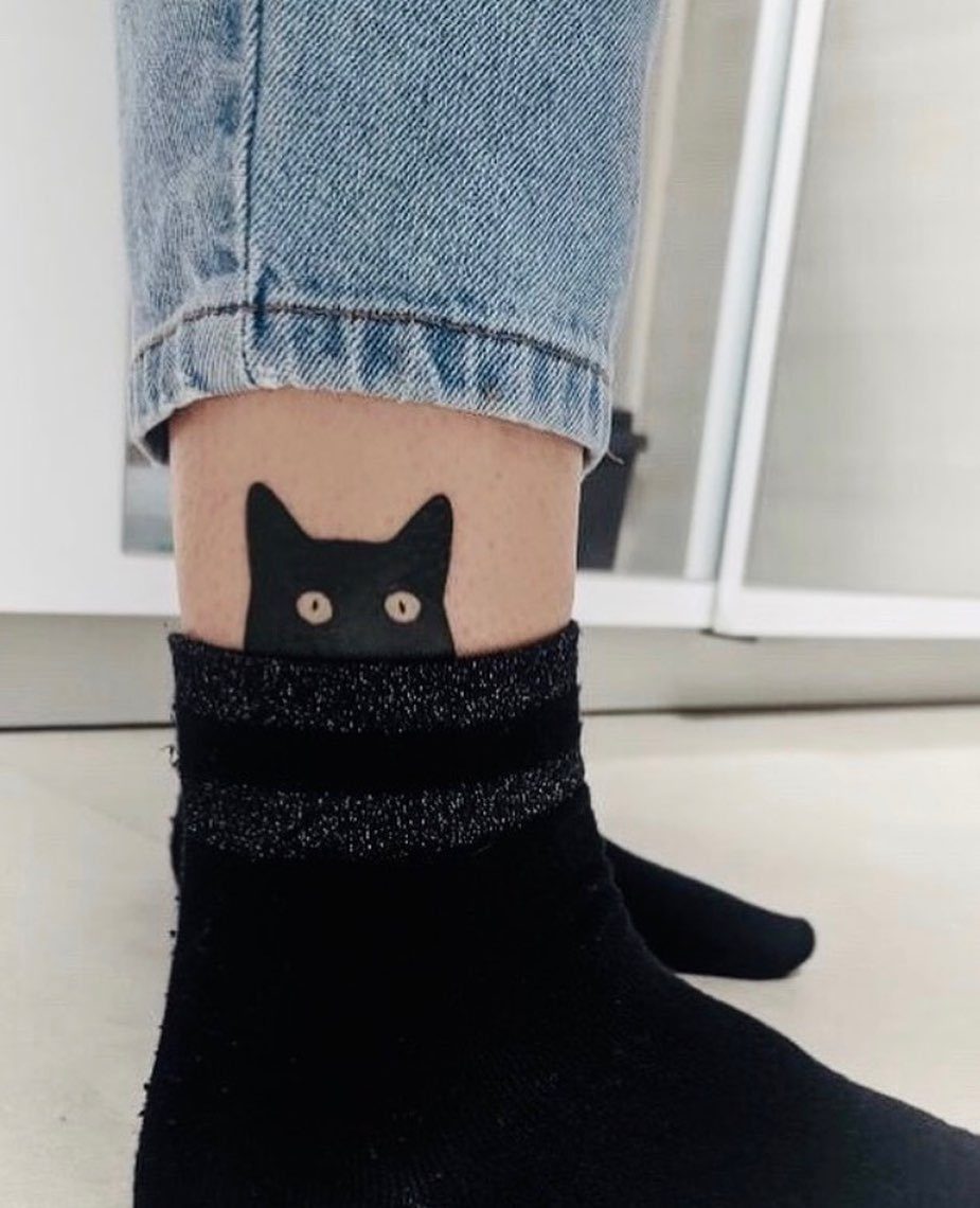 A black cat face tattoo with yellow eyes on a person's ankle above a black sock.