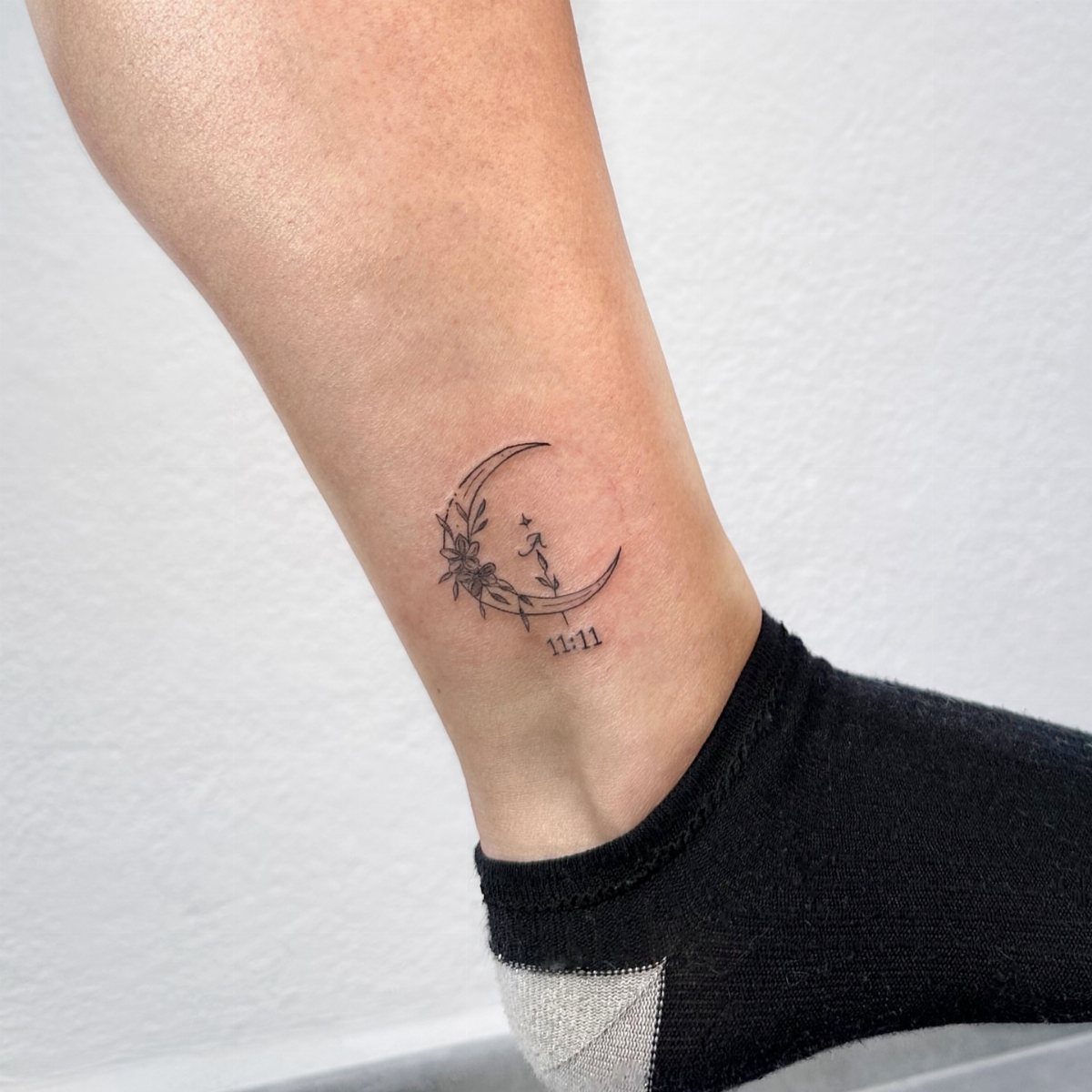 A delicate crescent moon tattoo with floral accents on a person's ankle.