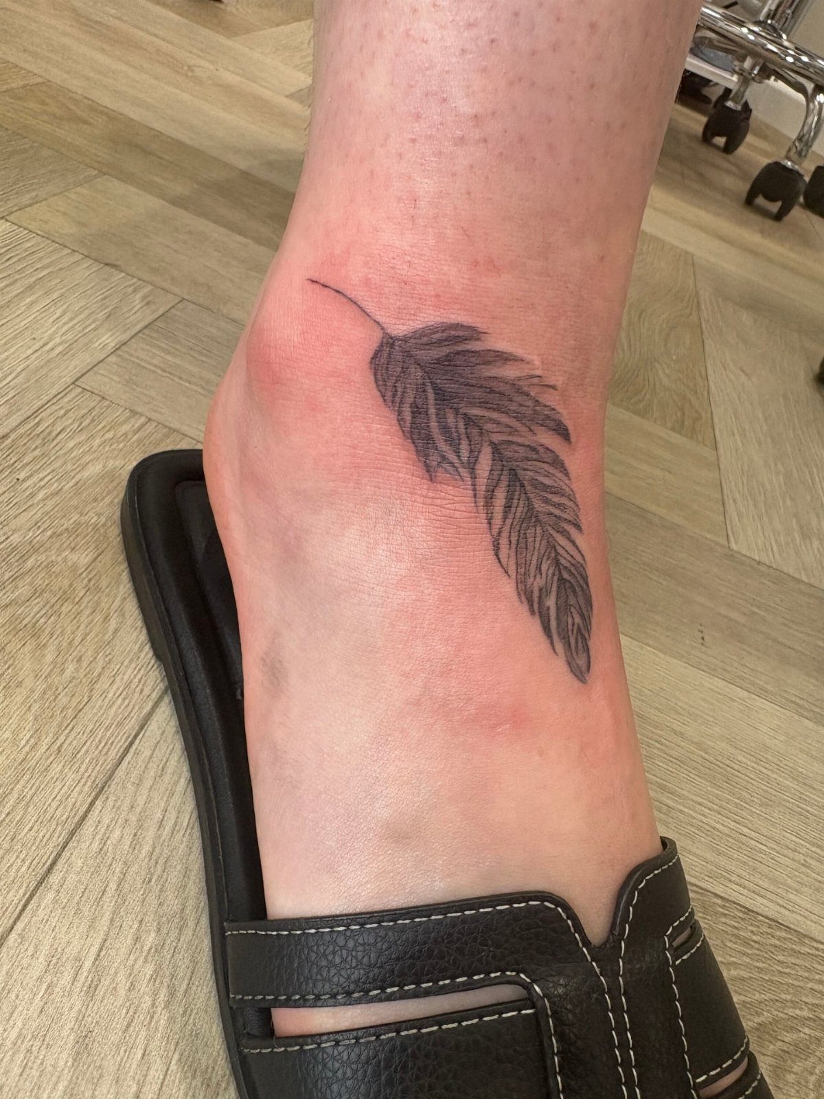 A detailed black ink feather tattoo on a person's ankle near a black sandal.