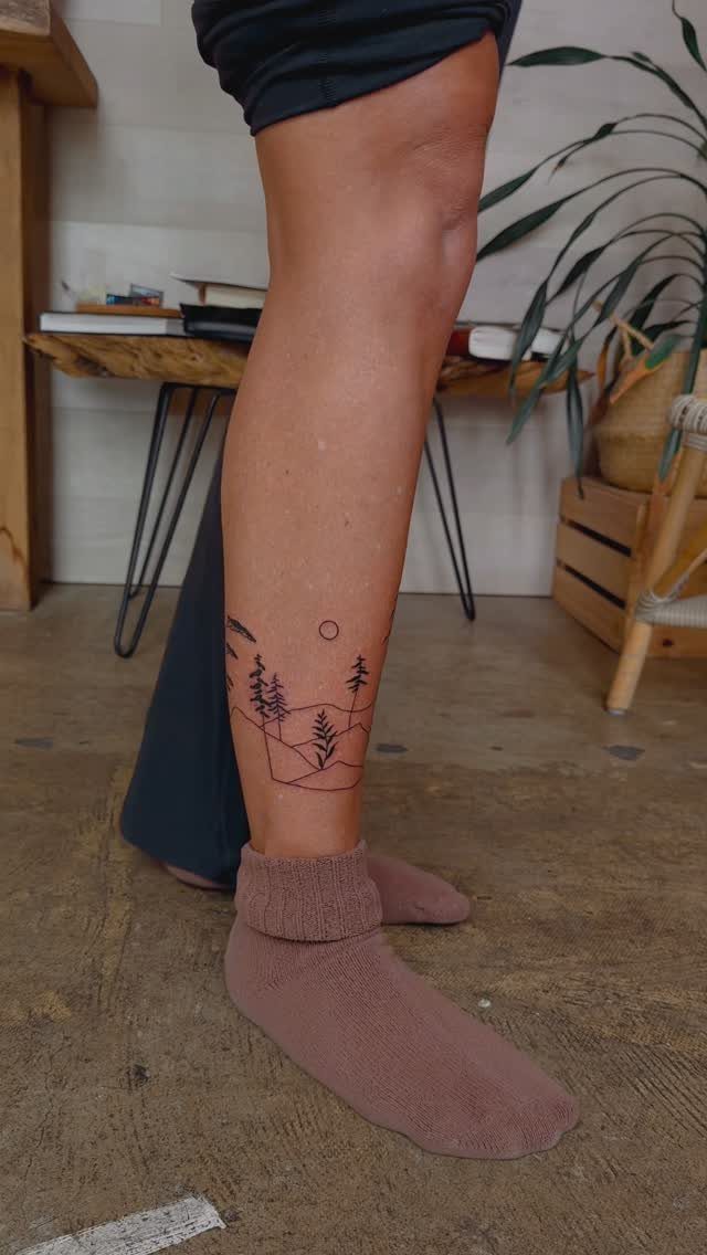 A person's lower leg displaying a minimalist line work landscape tattoo with pine trees and mountains.