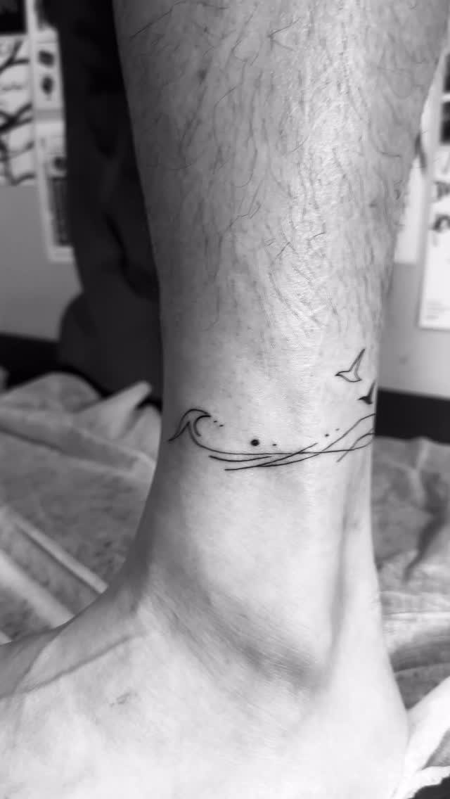 A minimalist line art tattoo of a small ocean wave and birds on a person's ankle.