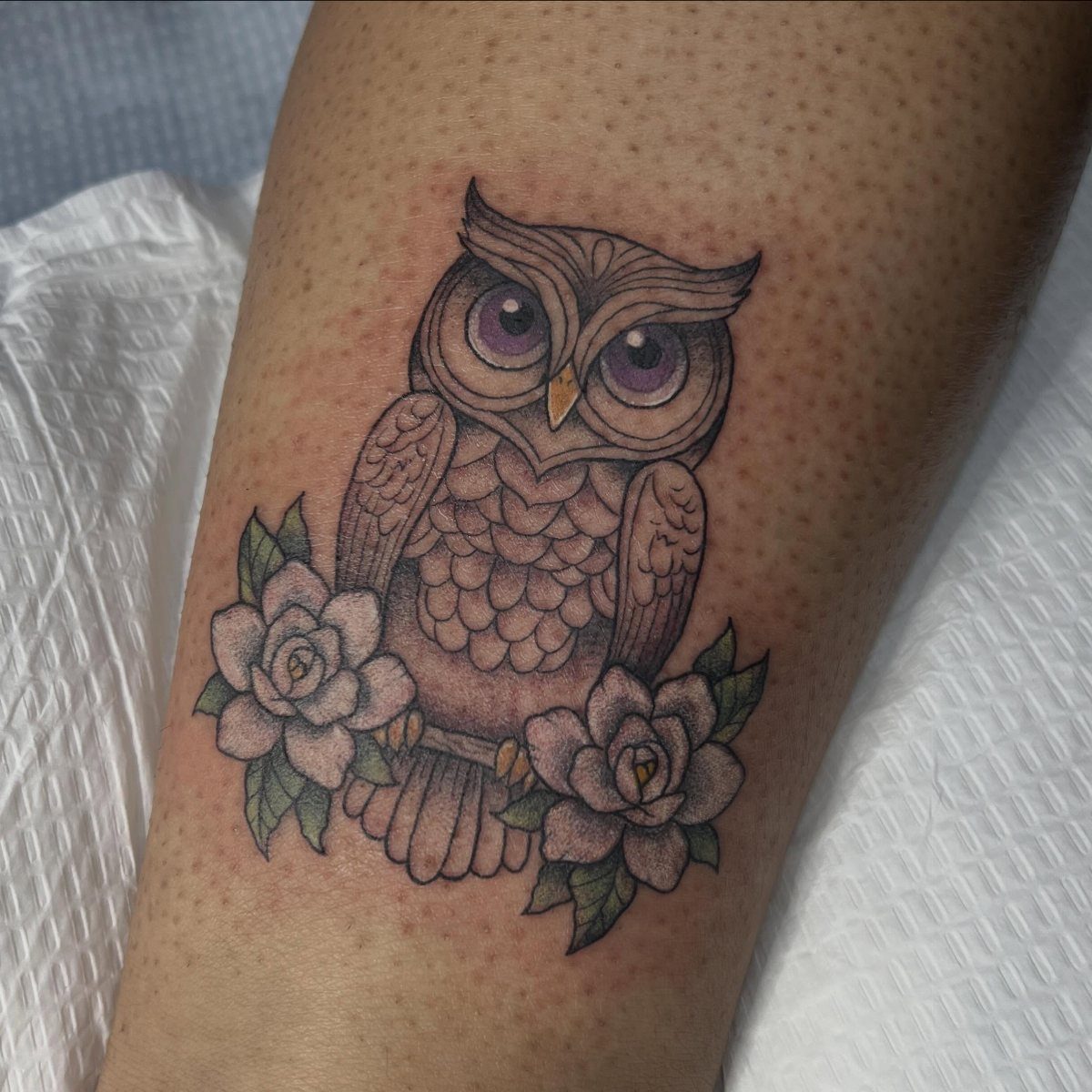 An owl tattoo with purple eyes surrounded by white roses on a person's skin.