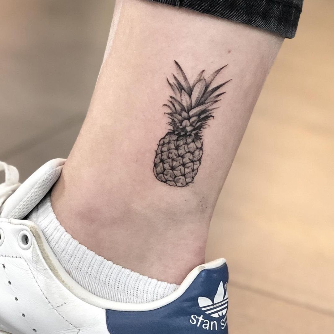 A small black ink pineapple tattoo located on a person's ankle near a white sneaker.