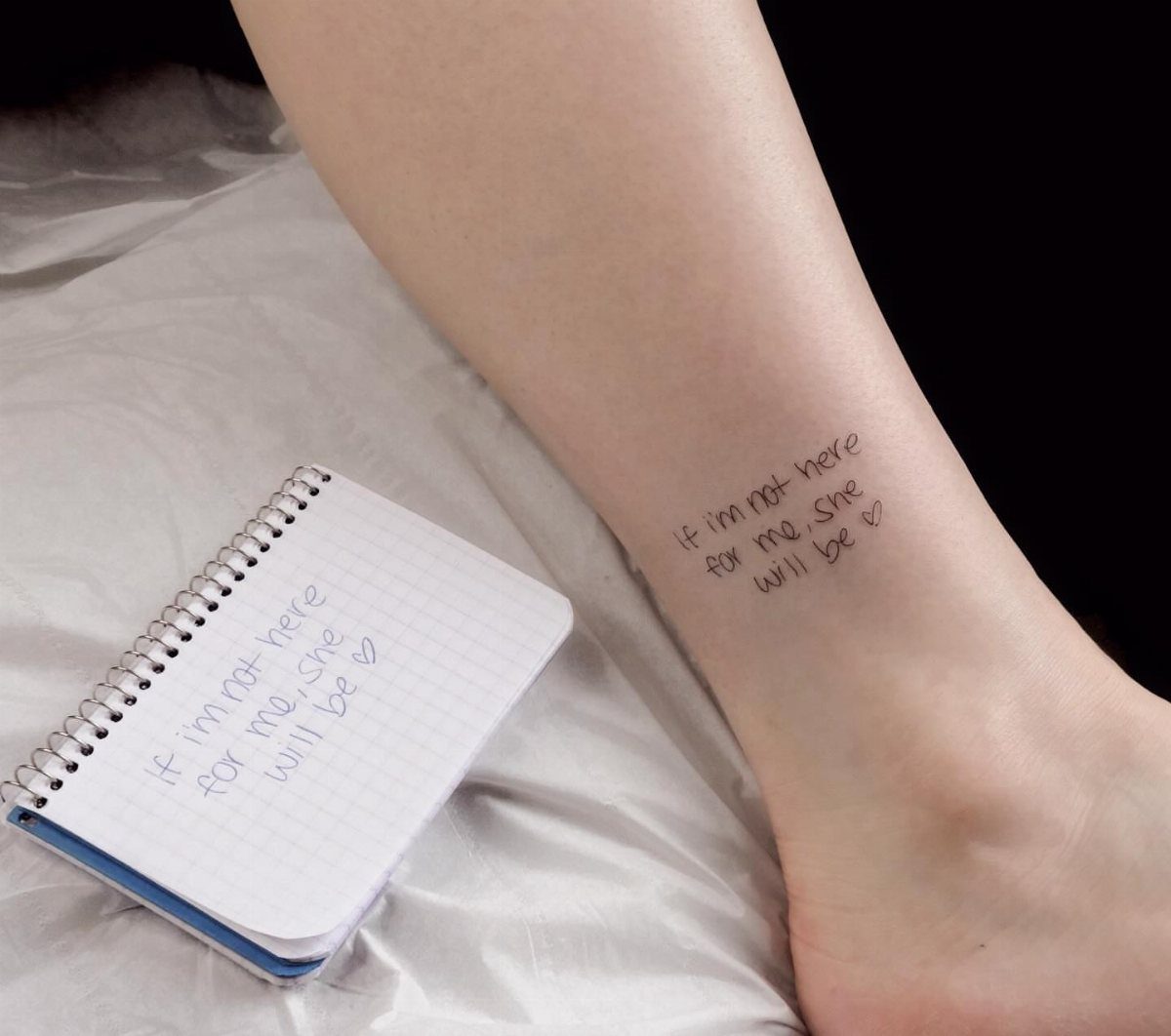 A person's ankle with a small handwritten quote tattoo next to a notebook.