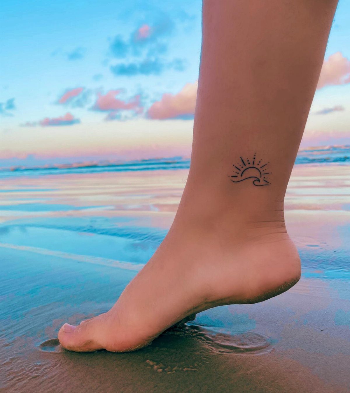 A minimalist sun and wave tattoo on a person's ankle at a beach sunset.