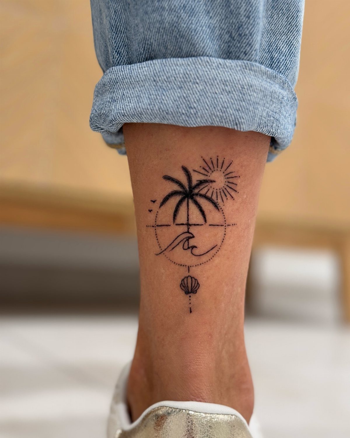 A fine line ankle tattoo featuring a palm tree, sun, wave, and seashell design.