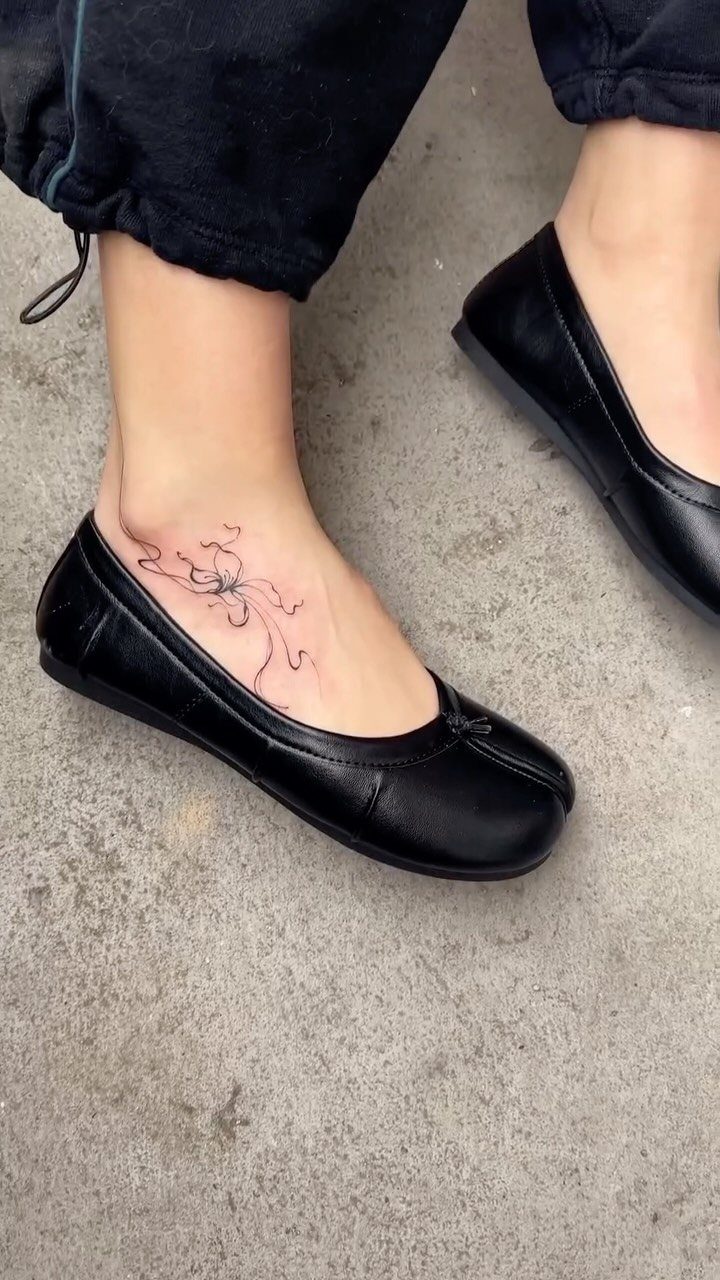 A delicate black ink floral tattoo placed on a person's ankle near black ballet flats.