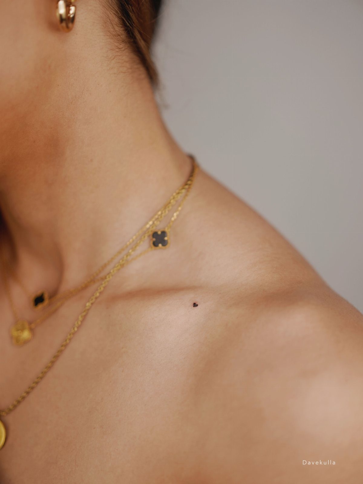 A tiny dark tattoo placed delicately on a person's collarbone near gold necklaces.