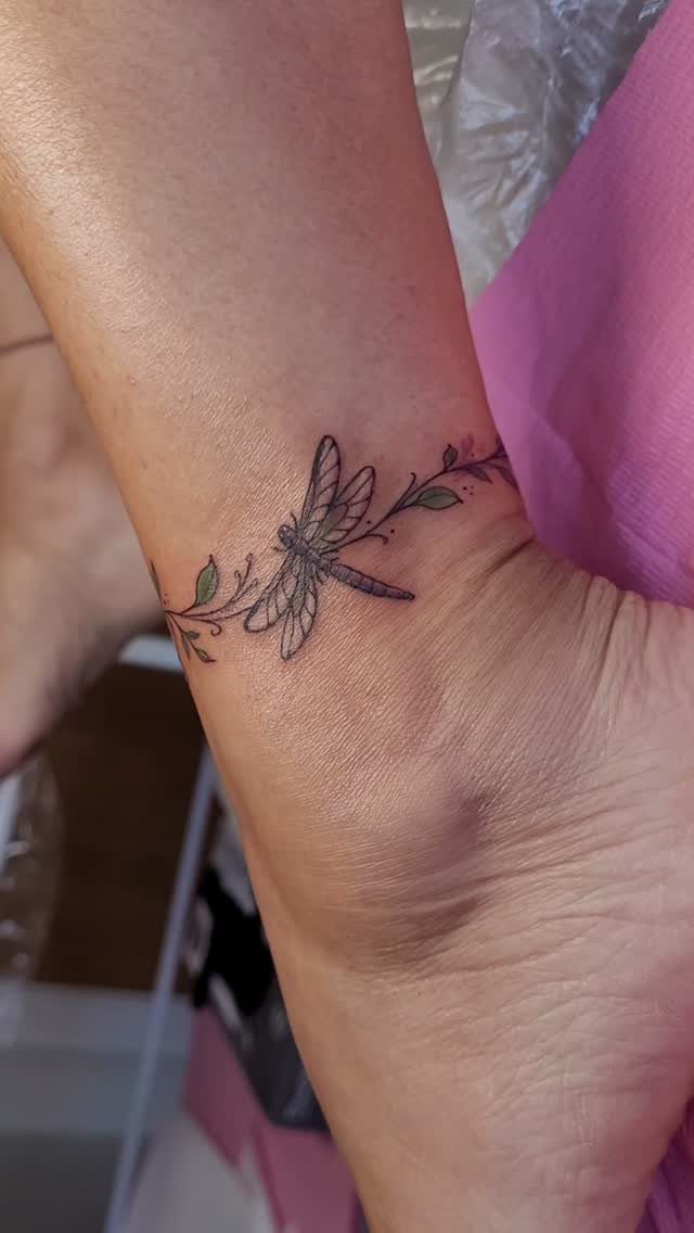 7 Feminine Ankle Tattoos for Every Style 6 A delicate dragonfly tattoo with fine lines and small leaves encircling a person's ankle.