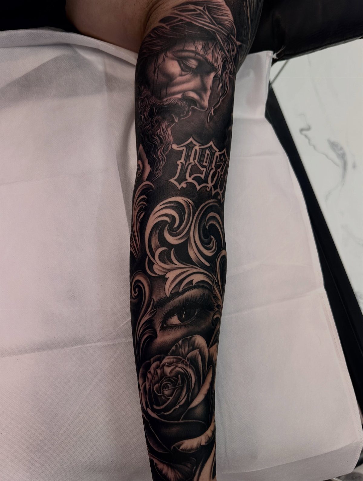 A detailed black and grey tattoo sleeve featuring a religious portrait, filigree, and a rose.
