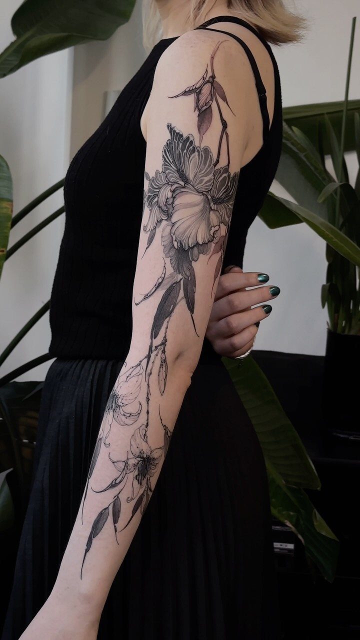 A woman's arm displaying a large fine line floral sleeve tattoo in black ink.