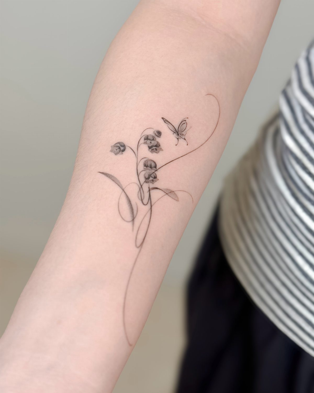 Fine line black ink tattoo of delicate flowers and a butterfly on a person's forearm.