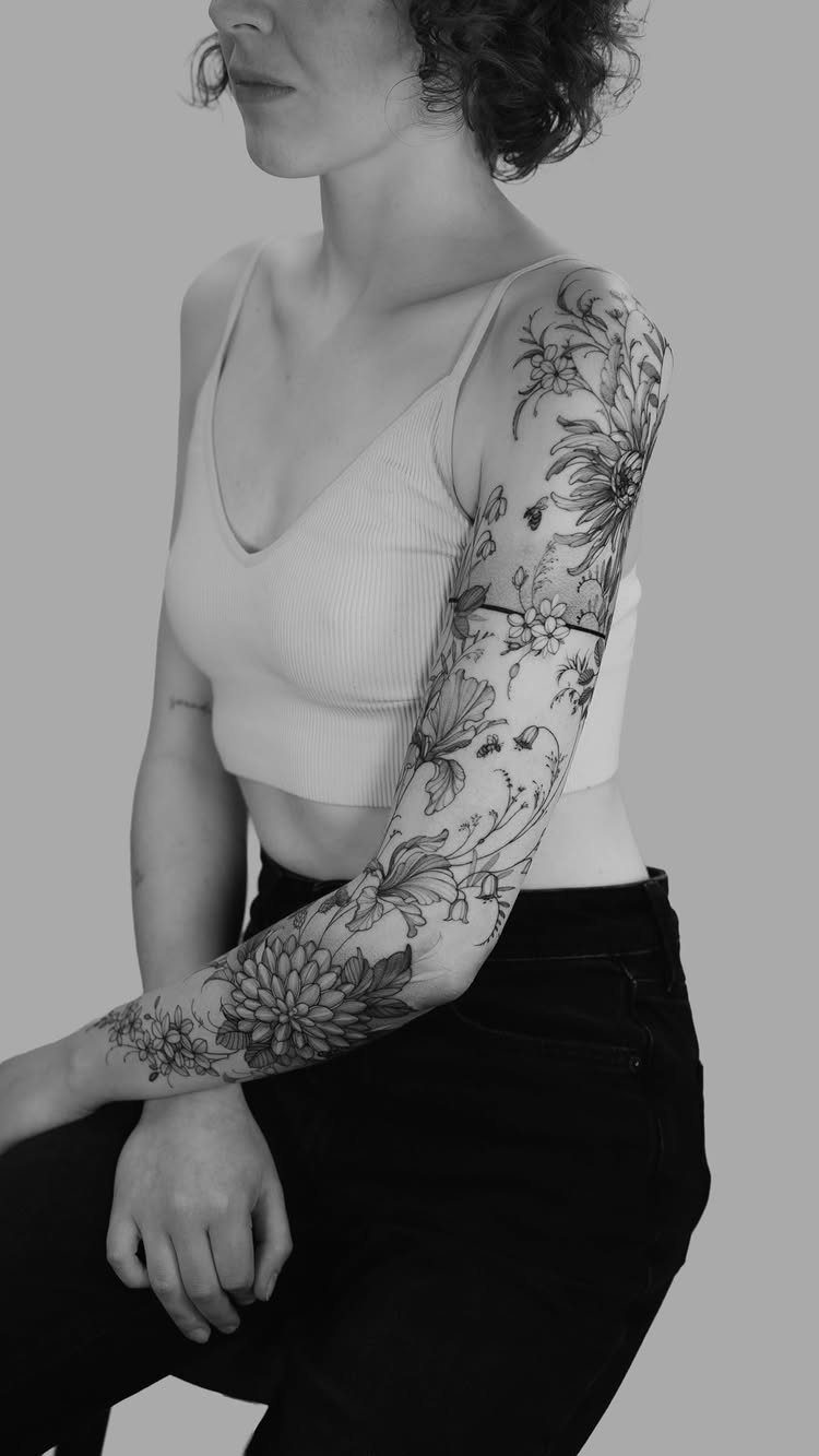 A woman's arm displaying a detailed black ink floral sleeve tattoo from shoulder to wrist.