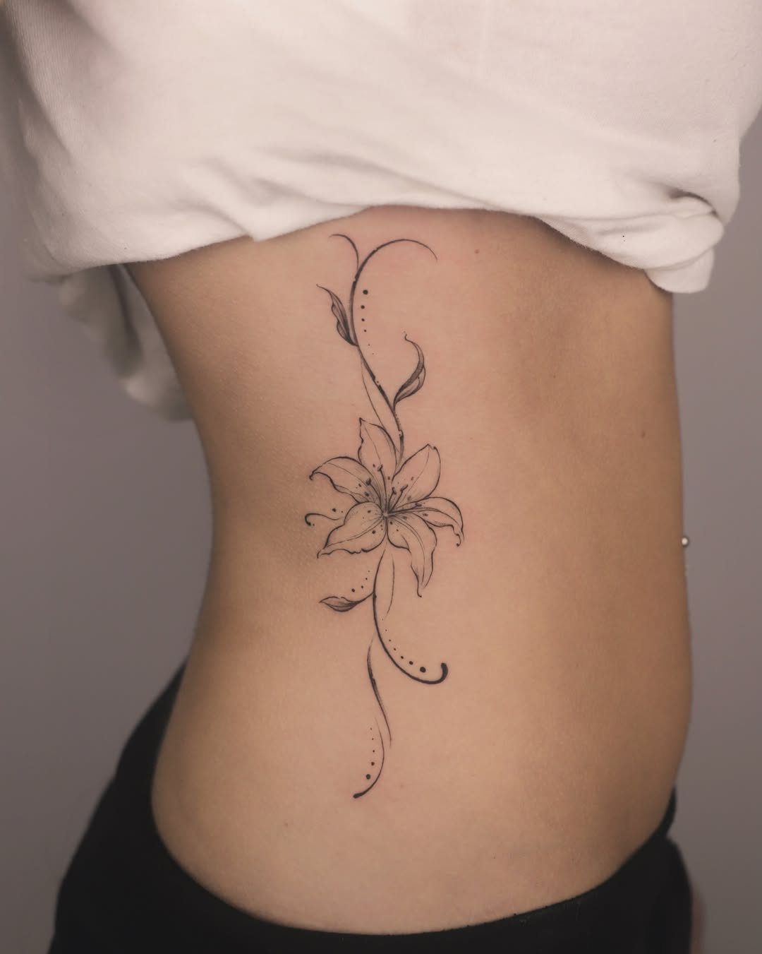 5 Flower Rib Tattoo Ideas for Women and Men 3 A black ink lily flower tattoo with delicate swirling vines on a person's ribs.
