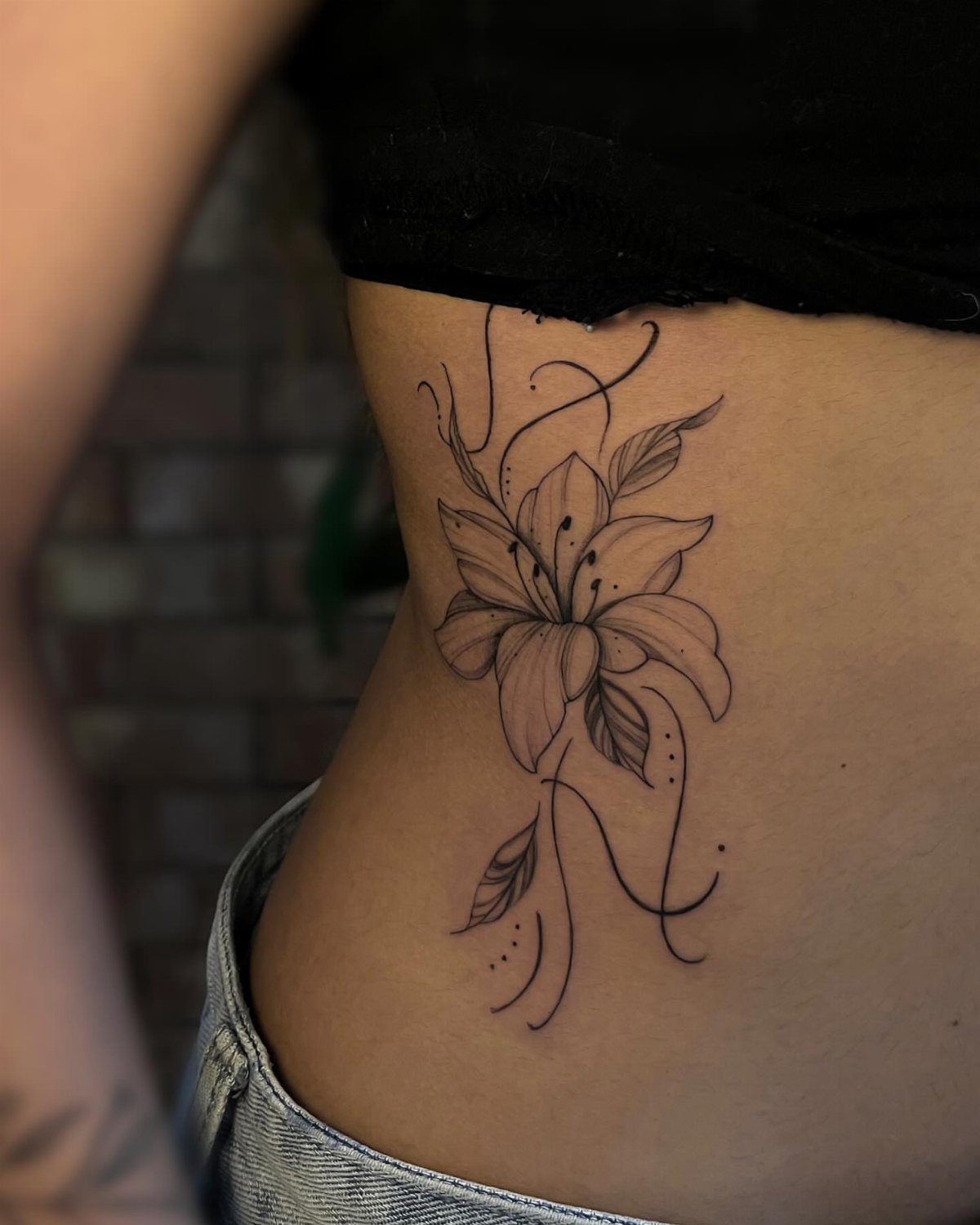 5 Flower Rib Tattoo Ideas for Women and Men 4 A black ink lily flower tattoo with swirling lines on a person's ribcage.