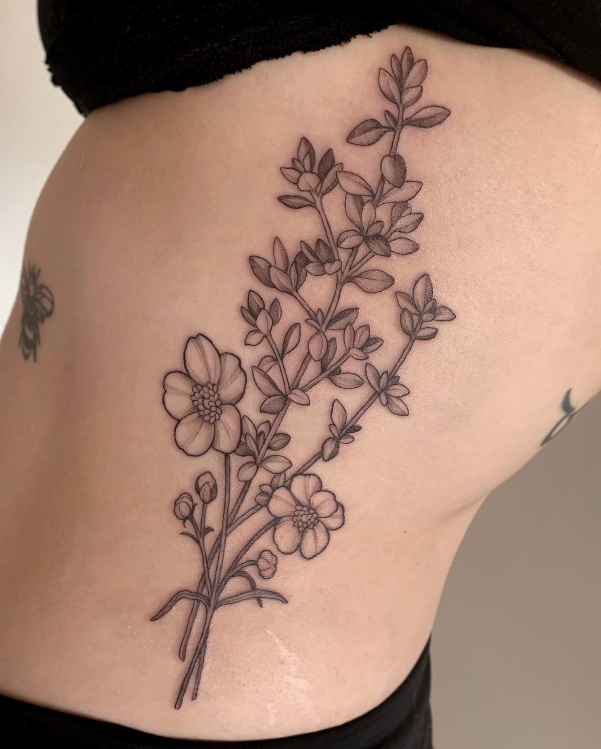 5 Flower Rib Tattoo Ideas for Women and Men 1 A black ink vertical wildflower stem tattoo with small blossoms on a person's ribs.