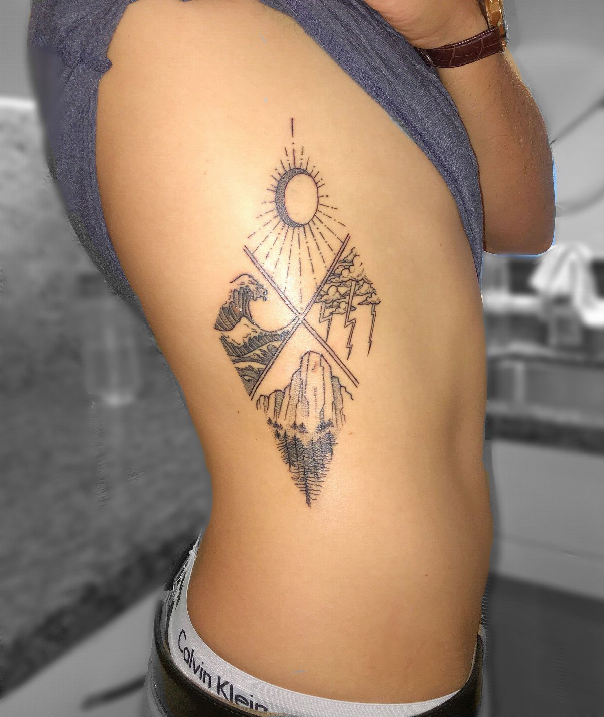 Four Elements Tattoo Design: 7 Styles That Hold Their Own 6 A fine line elemental tattoo on a person's ribs featuring a sun, waves, mountains, and lightning.
