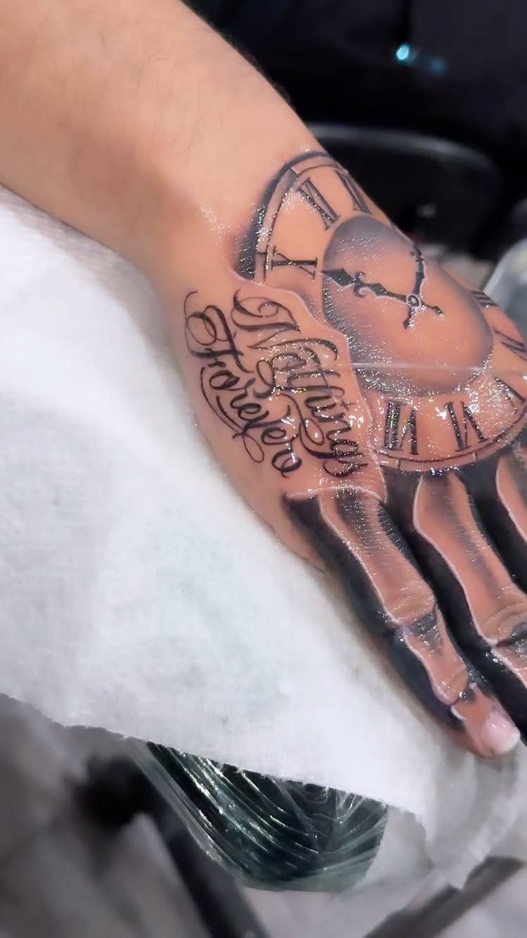 A hand with a large black and grey clock face tattoo and script lettering.