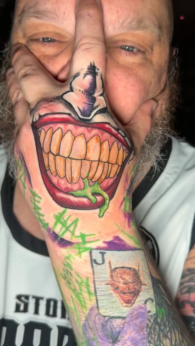 A man's hand and forearm displaying a large, colorful Joker-themed tattoo with a wide grin.