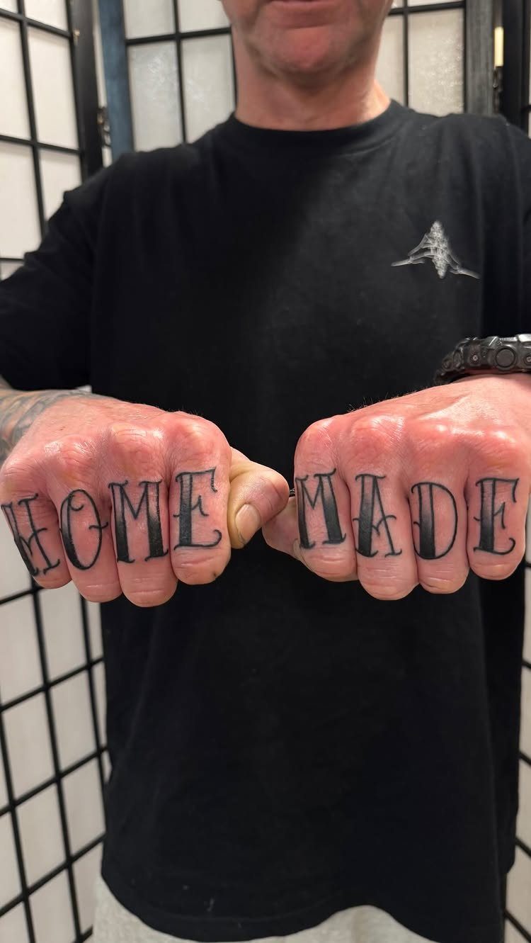 Man's clenched fists displaying black ink knuckle tattoos spelling out the words HOME MADE.