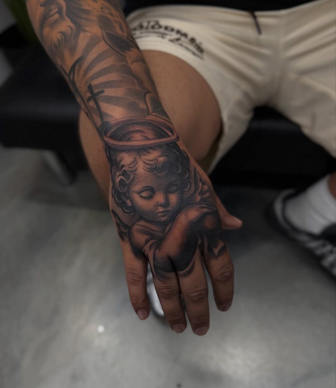 A man's hand and forearm featuring detailed black and grey realism tattoos of a cherub and clouds.