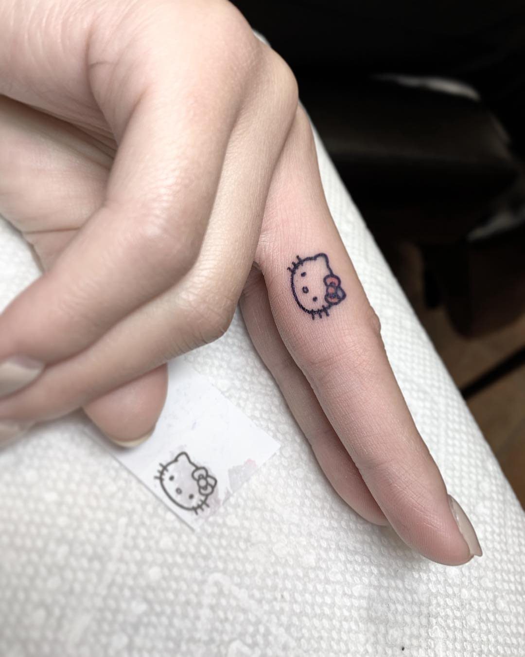A small Hello Kitty line art tattoo with a pink bow on a person's index finger.