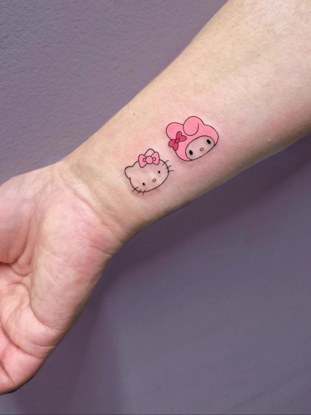 Two small Hello Kitty tattoos on a person's inner wrist against a purple background.