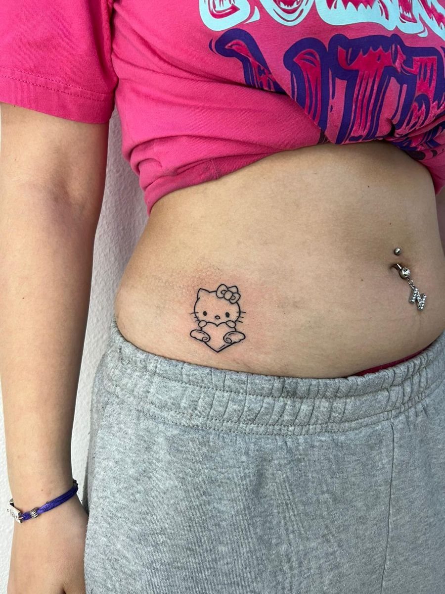 A small Hello Kitty tattoo on a person's lower abdomen near their navel piercing.