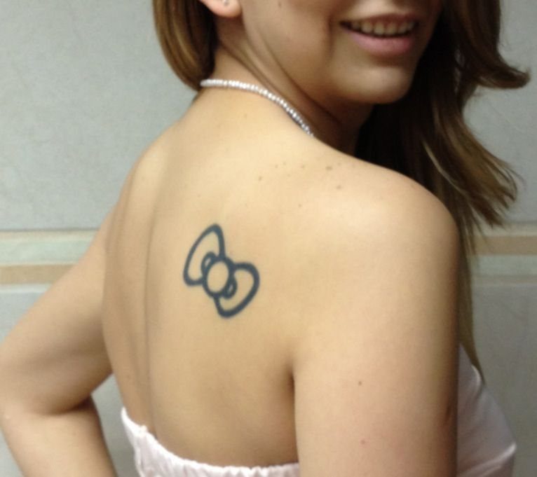 A small black bow outline tattoo located on a person's upper back and shoulder.