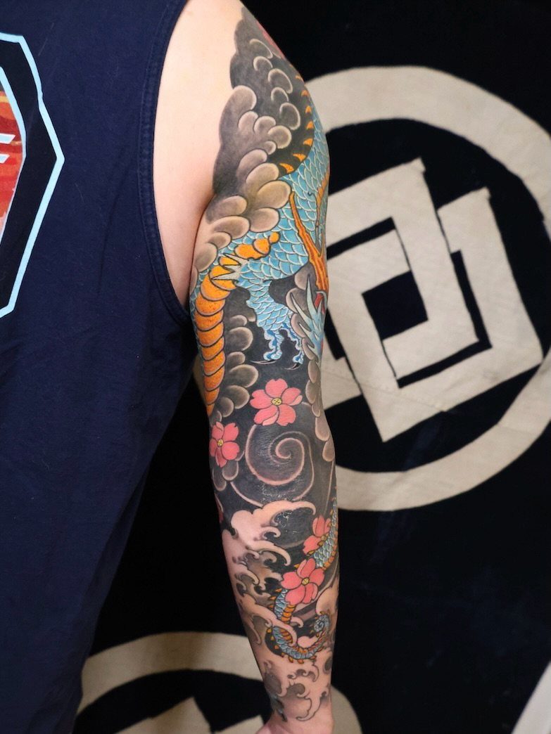 Japanese Dragon Tattoos: 19 Designs With Meaning and Style 3 A person's arm displaying a colorful Japanese sleeve tattoo with a dragon and cherry blossoms.