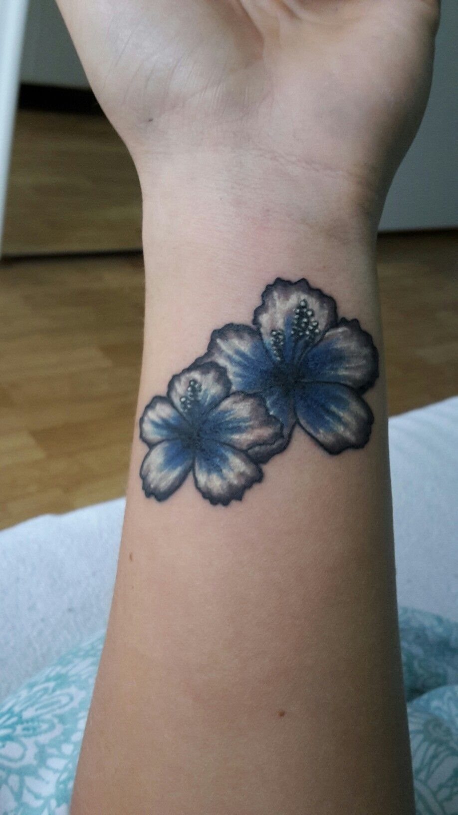 Two blue hibiscus flowers tattooed on a person's wrist with dark shading and white highlights.
